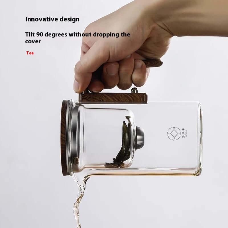 Hand holding a glass teapot with a wooden handle, pouring tea while tilting it at a 90° angle.
